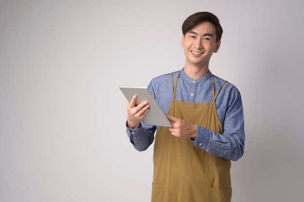 Portrait of young asian man wearing apron holding tablet over white background