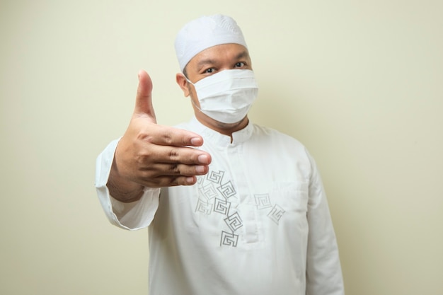 portrait of young asian muslim man wearing mask he is offering handshake and smiling