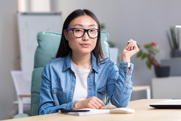 Portrait of a young asian student girl in glasses and denim clothes sitting at a desk with a