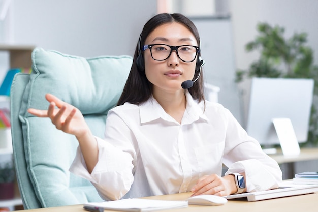 Portrait of a young asian woman in glasses and headphones with a microphone sitting in the office