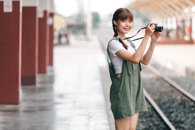 Portrait Young asian woman smiling tourist Traveler girl walking and with a holding the camera waits train travel journey is taken in railway platform Thailand summer relax vacation Concept