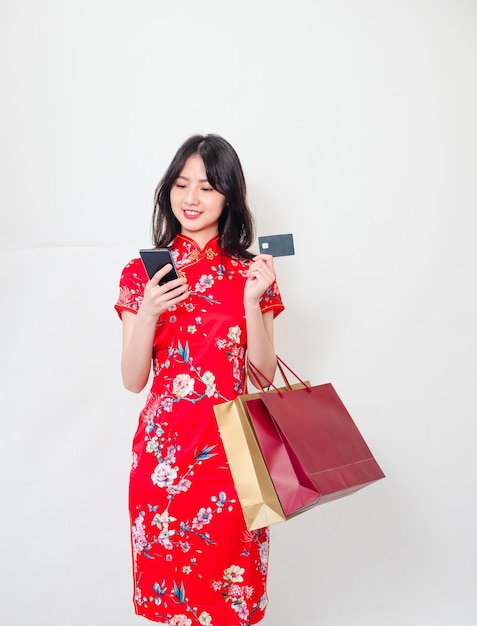 Portrait of a young asian woman wearing traditional cheongsam qipao dress holding shopping bagssmartphone and showing credit card isolated over white background