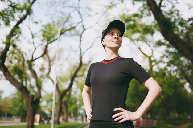 Portrait of young athletic beautiful brunette woman in black uniform and cap standing, resting and looking aside after running, training in city park outdoors