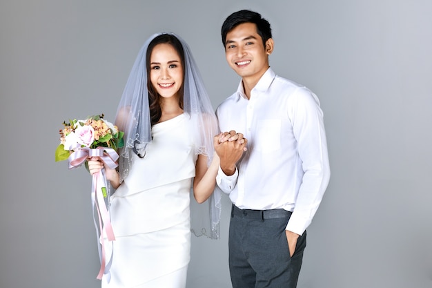 Portrait of young attractive Asian couple holding hands, man wearing white shirt, woman wearing wedding gown with veil holding bouquet of flower. Concept for pre wedding photography.