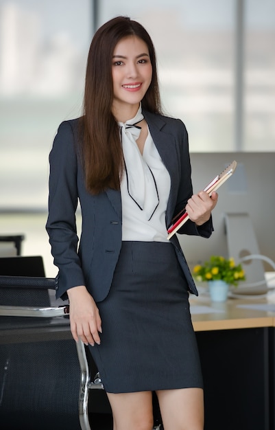 Portrait of young attractive Asian woman in black business suit standing holding notebook in modern looking office with blurry windows background. Concept for modern office lifestyle.