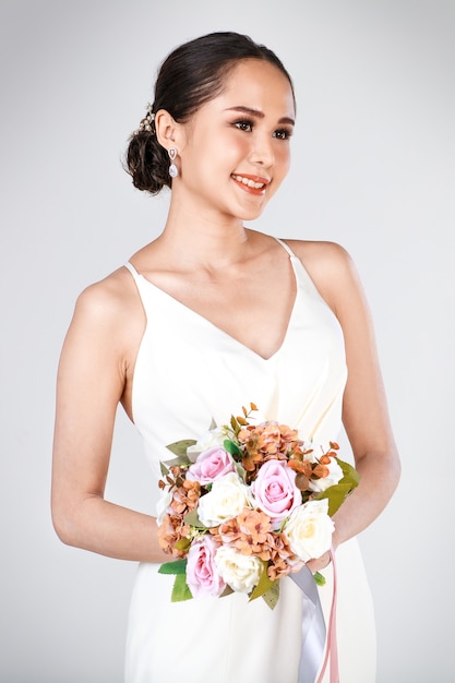 Portrait of young attractive Asian woman wearing white wedding dress smiling holding bouquet of flower. Concept for pre wedding photography.