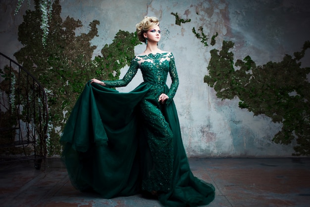 Portrait of young attractive blonde woman in a beautiful green dress. Textured background, interior. Luxury hairstyle