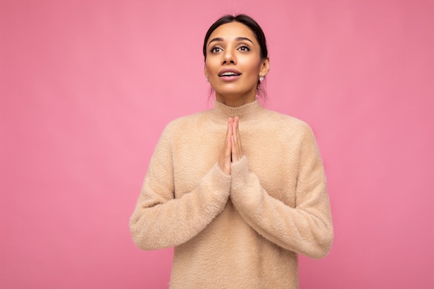 Portrait of young attractive brunette woman with sincere emotions wearing beige pullover isolated over pink background with copy space and keeping hand in praying gesture believing in good luck