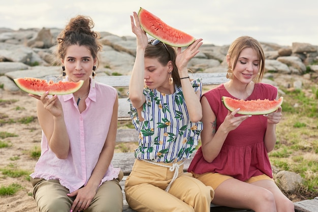 Portrait of young attractive Caucasian women holding slices of watermelon and posing on camera while sitting on bench outdoors