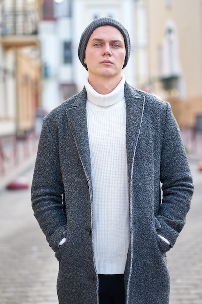 Portrait of a young attractive hipster man wearing a gray coat, white sweater and black jeans