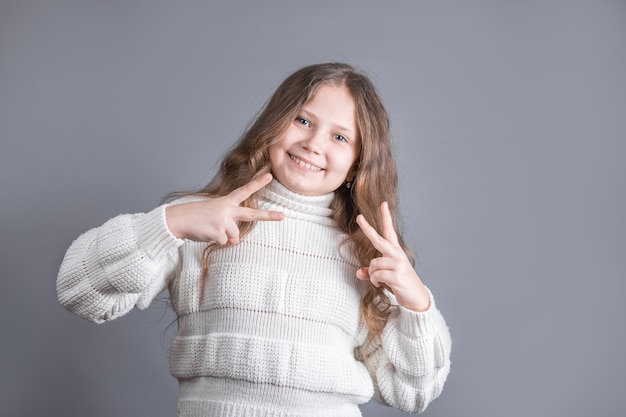 Portrait of a young attractive little girl with blond hair in sweater showing v-sign,peace sign, victory gesture on both hands on a gray studio background.