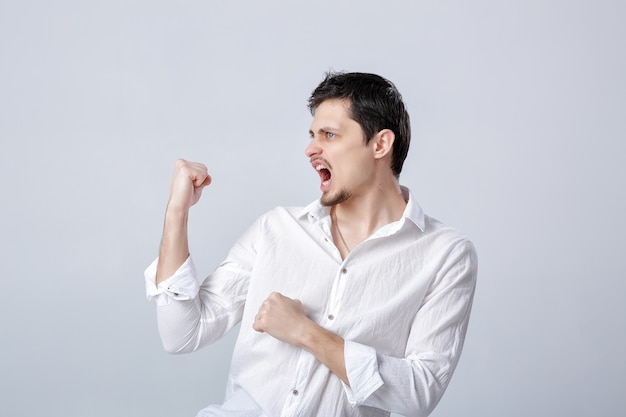 Portrait of young attractive man with dark hair in a white shirt raises his hands above his head on the grey background. the gesture of a winner