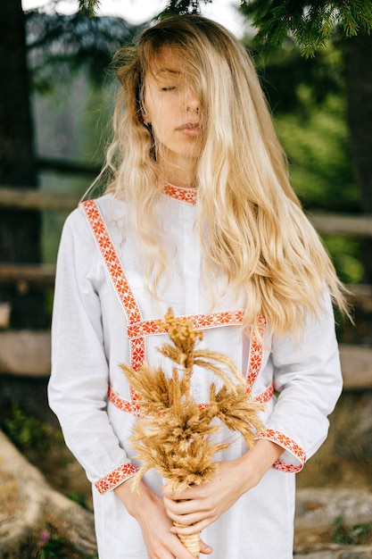 Portrait of young attractive sensitive blonde girl in white dress with ornament posing with spikelets bouquet over nature