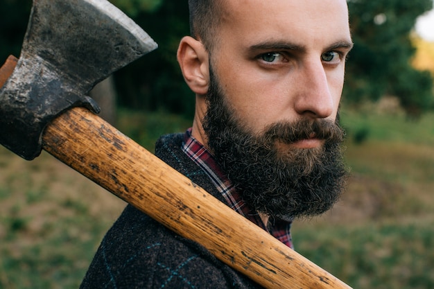 Portrait of young bearded hipster logger with ax in forest