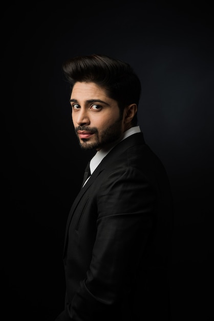 Portrait of young bearded Indian businessman against black background, moody lighting