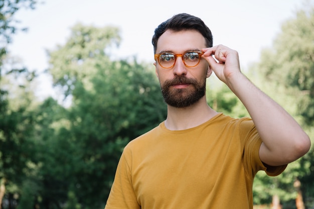 Portrait of young bearded man wearing stylish eyeglasses, looking at camera, standing outdoors