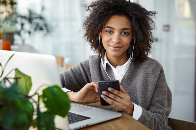 Portrait of young beautiful african american woman using laptop and cellphone while sitting by window in cafe