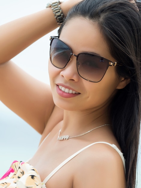 Portrait of young beautiful asian woman wearing sunglasses and standing on the beach. Girl touching her hair. Summer time, relaxation or vacations concept