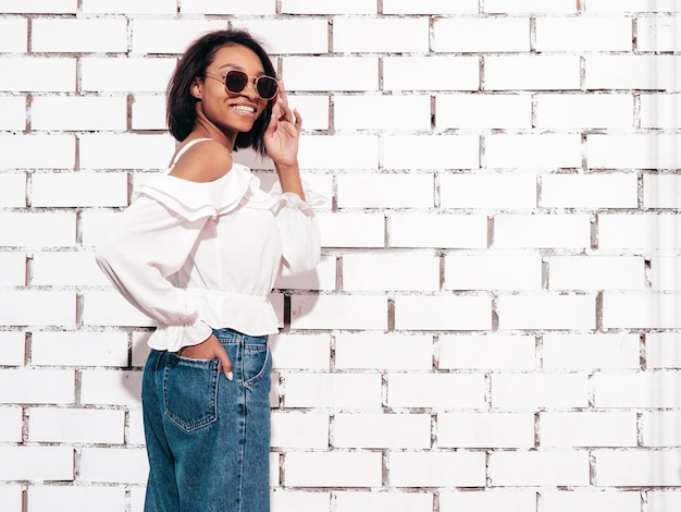 Portrait of young beautiful black woman Smiling model dressed in summer jeans clothes Sexy carefree female posing near white brick wall in studio Tanned and cheerful