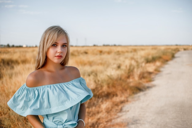 Portrait of a young beautiful caucasian blonde girl in a light blue dress standing on a field with sun-dried grass next to a small country road