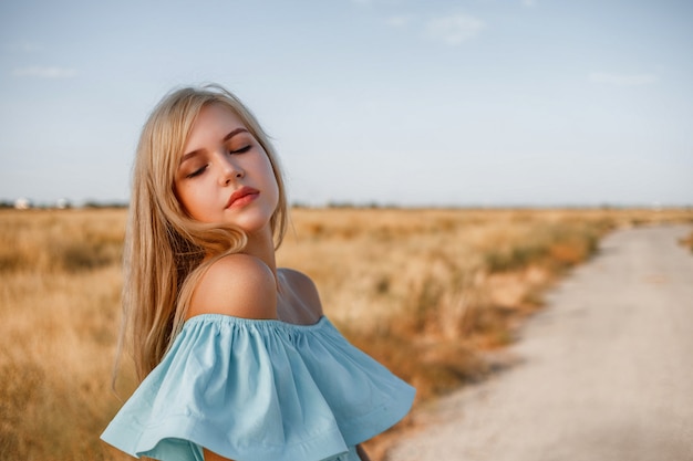 Portrait of a young beautiful caucasian blonde girl in a light blue dress standing on a field with sun-dried grass next to a small country road