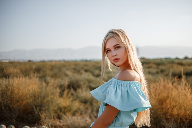 Portrait of a young beautiful caucasian blonde girl in a light blue dress standing on a watermelon field with sun-dried grass during sunset