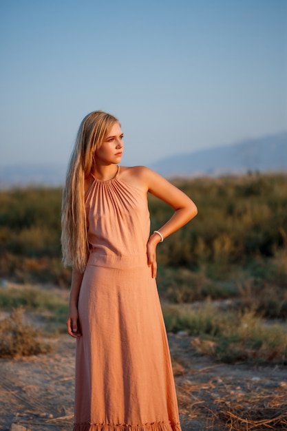 Photo portrait of a young beautiful caucasian blonde woman in a pink dress in a deserted field against the sunset