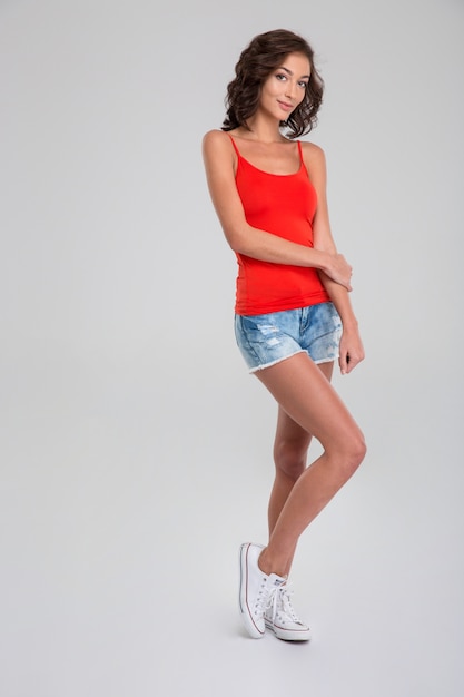 Photo portrait of young beautiful curly woman in red top, jeans shorts and white sneakers standing on white wall