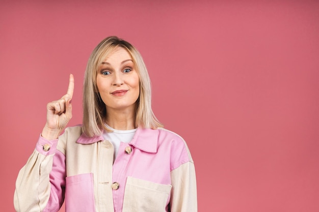 Portrait of a young beautiful cute cheerful blonde woman smiling looking at the camera isolated over pink background. Pointing finger up.