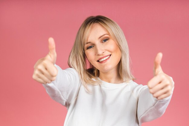 Photo portrait of a young beautiful cute cheerful blonde woman smiling looking at the camera isolated over pink background. thumb up sign.