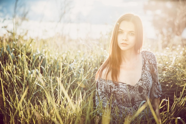 Portrait of a young beautiful girl in a dress sitting in the grass during sunset