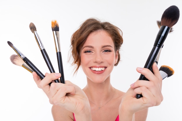 Portrait of young beautiful girl show her makeup brushes isolated on white wall