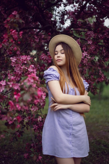 Portrait young beautiful girl teenager in a purple dress and in a hat stands by a blooming pink apple tree