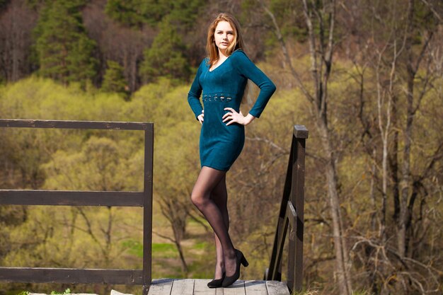 Photo portrait of a young beautiful girl in a tight short dark turquoise dress posing against a background of a spring park