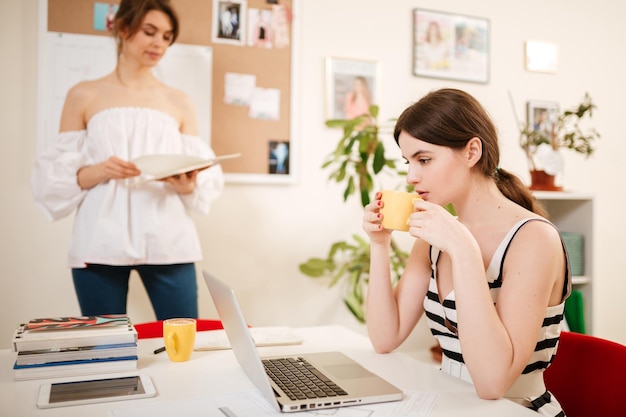 Portrait of young beautiful girl with dark hair sitting at the desk and drinking coffee with laptop on table while lady standing and reading book nearby in office