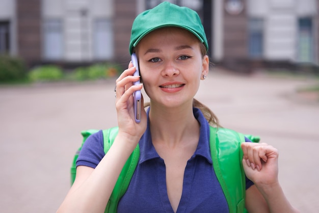 Portrait of young beautiful happy courier teenager girl food delivery woman with green thermo box for food delivering food outdoors in cap and uniform using cell mobile phone calling on smartphone