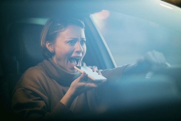 Portrait of a young beautiful happy woman who is driving a car, talking at the smartphone and arguing with someone.