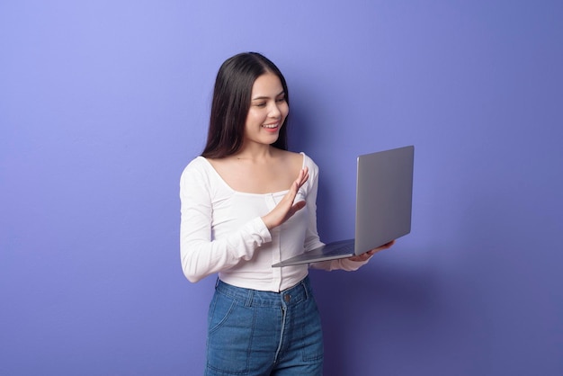 A portrait of young beautiful smiling woman is holding laptop over isolated purple background studio