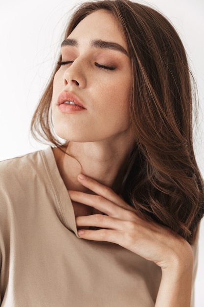 Portrait of young beautiful woman posing indoors isolated on white wall touching neck with eyes closed.