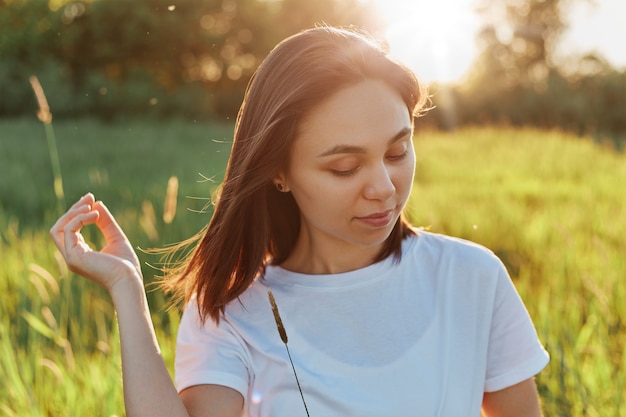 Portrait of young beautiful woman wearing white casual T-shirt with dark hair posing on sunset in field or meadow, looking away with dreamy facial expression, enjoying sunset and nature.