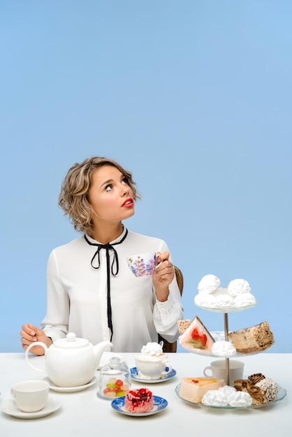 Portrait of young beautiful woman in white blouse sitting at table with sweets, holding cup over blue wall