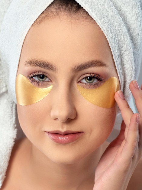 Portrait of young beautiful woman with towel on her hair applying golden hygrogel eye patches under her eyes Skin treatment and morning routine concept