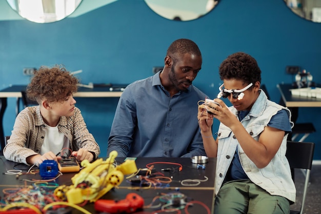 Portrait of young black boy building robots in engineering class with male teacher helping