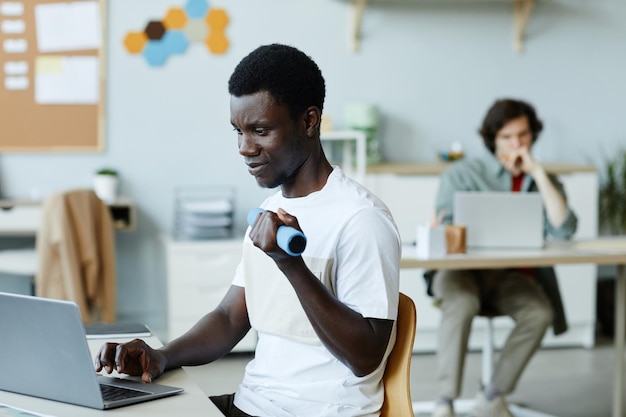 Portrait of young black man holding dumbbell in hand while using computer at workplace office fitnes