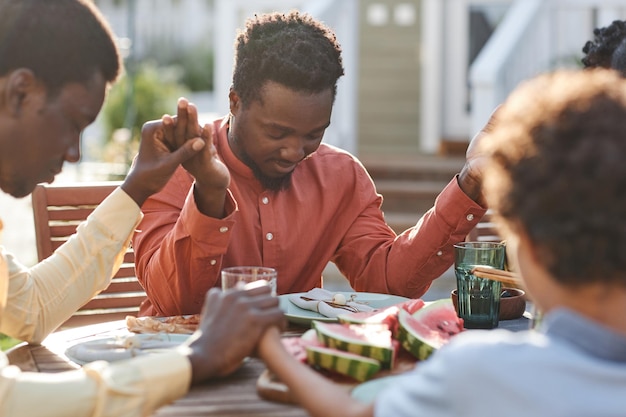 Portrait of young black man saying grace at table outdoors during family gathering and holding hands