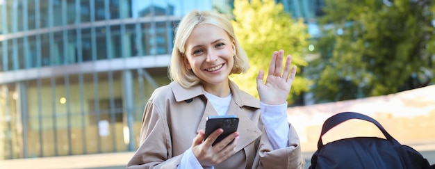 Portrait of young blonde college girl student sitting on street bench holding smartphone seeing a