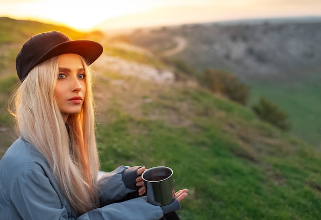 Portrait of young blonde girl with black cap, holding steel mug with hot tea on of sunset with copy space.