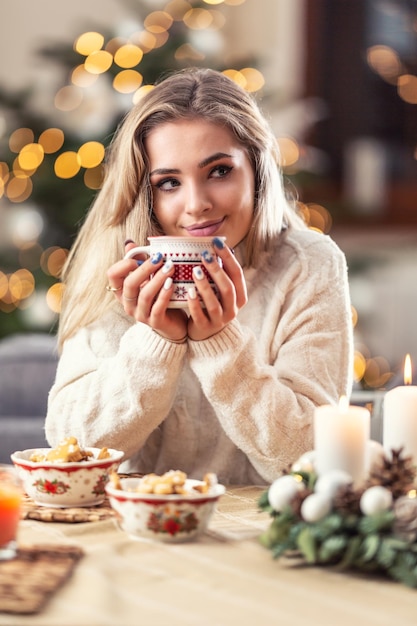 Portrait of a young blonde sitting at a Christmas table in front of a Christmas tree drinking hot coffee tea or punch