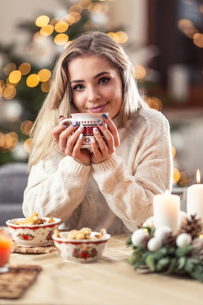 Portrait of a young blonde sitting at a Christmas table in front of a Christmas tree drinking hot coffee tea or punch