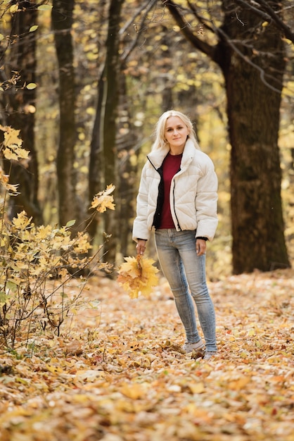 Portrait of a young blonde woman in the autumn forest, with a bouquet of yellow leaves in her hands.